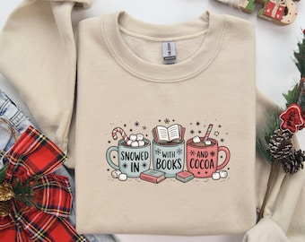 Könnte beinhalten: Beiges Sweatshirt mit einer Grafik von drei Tassen mit dem Text "Snowed in with books and cocoa". Die Tassen sind blau, grau und rot und enthalten Marshmallows, ein Buch und Kakao. Das Sweatshirt hat einen Rundhalsausschnitt und lange Ärmel.