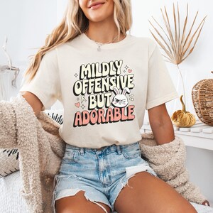 K&ouml;nnte beinhalten: Ein wei&szlig;es T-Shirt mit einer pinkfarbenen und gelben Grafik, auf der "Mildly Offensive But Adorable" steht, mit einem Comic-Kaninchen mit w&uuml;tendem Gesichtsausdruck.