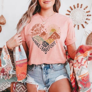 T-shirt with a heart-shaped mountain and floral design, sun rising in the background. Text reads: Where the Wild Things Hike Barefoot. The main colors are orange, green, red and yellow tones.