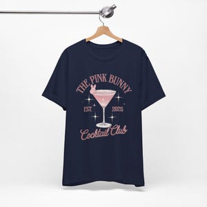A trendy cocktail-themed t-shirt featuring a pink martini glass with a bunny accent and vintage lettering, perfect for bar lovers and aesthetic outfits. The main colors are pink and white.