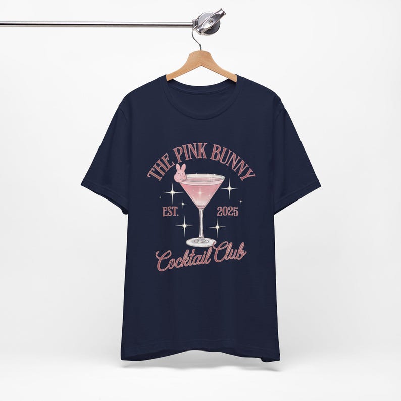 A trendy cocktail-themed t-shirt featuring a pink martini glass with a bunny accent and vintage lettering, perfect for bar lovers and aesthetic outfits. The main colors are pink and white.