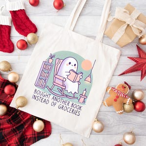 A natural-colored tote bag featuring a cute ghost reading a book with a humorous message about choosing books over groceries. The text says: Bought Another Book Instead Of Groceries. Main colors are blue, orange, white, pink and black.