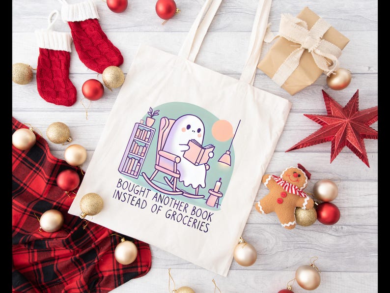 A natural-colored tote bag featuring a cute ghost reading a book with a humorous message about choosing books over groceries. The text says: Bought Another Book Instead Of Groceries. Main colors are blue, orange, white, pink and black.