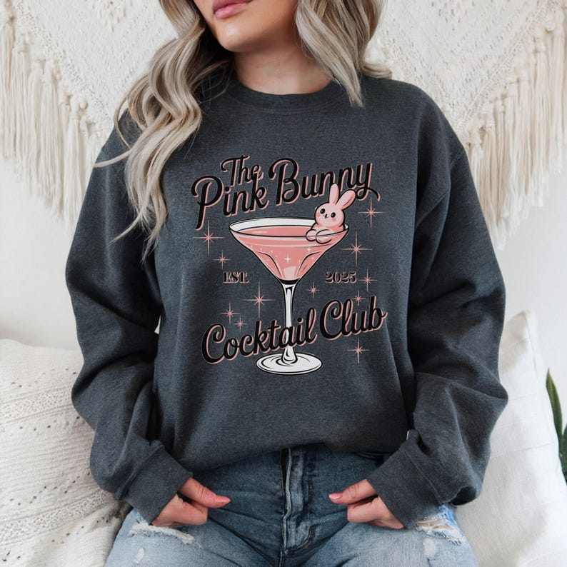 A sweatshirt featuring a pink martini glass with a bunny lounging on the rim, surrounded by vintage-style lettering that reads: The Pink Bunny Cocktail Club, Est. 2025. The main colors are pink and black.