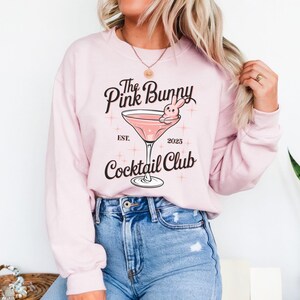 A sweatshirt featuring a pink martini glass with a bunny lounging on the rim, surrounded by vintage-style lettering that reads: The Pink Bunny Cocktail Club, Est. 2025. The main colors are pink and black.