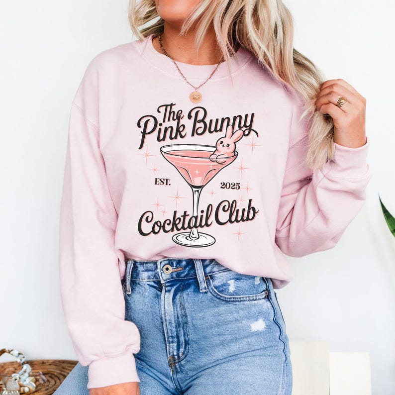 A sweatshirt featuring a pink martini glass with a bunny lounging on the rim, surrounded by vintage-style lettering that reads: The Pink Bunny Cocktail Club, Est. 2025. The main colors are pink and black.
