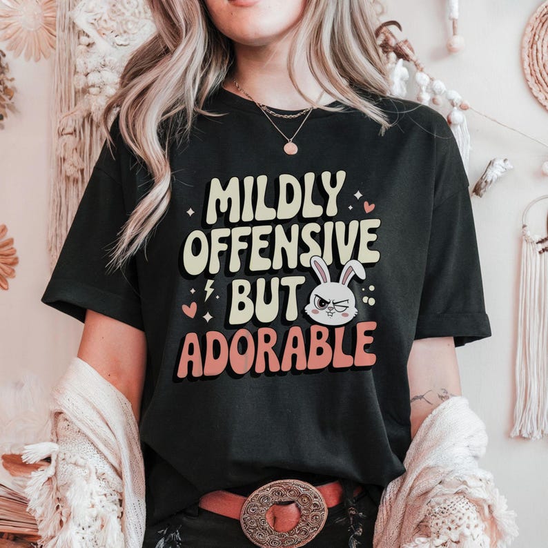 Funny graphic T-shirt featuring a sassy bunny with the phrase "Mildly Offensive But Adorable," perfect for humor-loving animal lovers. The main colors are white, beige and peach.