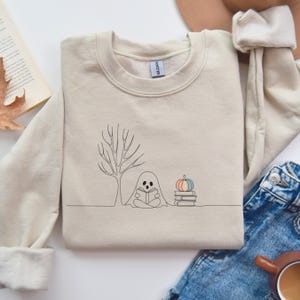 Può includere: Felpa girocollo beige con un disegno a tratto minimalista di un fantasma che legge un libro sotto un albero, con una zucca e dei libri. Il design è in nero. Grafica a tema autunnale.