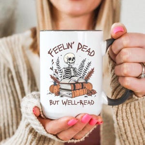 A white mug with a black or pink handle featuring a skeleton reading a book, surrounded by fall leaves and pumpkins, with the text: Feelin' Dead But Well-Read. Main colors are brown and orange.