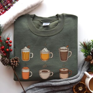 May include: An olive green sweatshirt featuring a graphic of six different hot beverages, including coffee and cocoa, each in a unique mug. The mugs are in shades of orange, brown, and white, with steam rising from each. A cozy, seasonal theme.