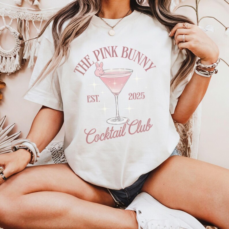 A trendy cocktail-themed t-shirt featuring a pink martini glass with a bunny accent and vintage lettering, perfect for bar lovers and aesthetic outfits. The main colors are pink and white.