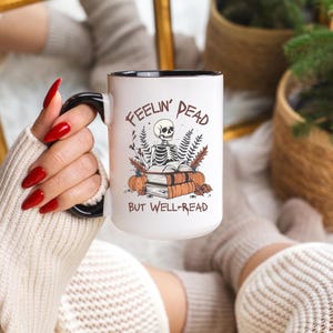 A white mug with a black or pink handle featuring a skeleton reading a book, surrounded by fall leaves and pumpkins, with the text: Feelin' Dead But Well-Read. Main colors are brown and orange.