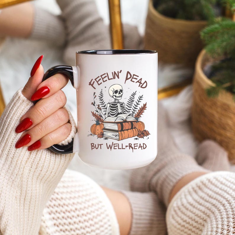 A white mug with a black or pink handle featuring a skeleton reading a book, surrounded by fall leaves and pumpkins, with the text: Feelin' Dead But Well-Read. Main colors are brown and orange.