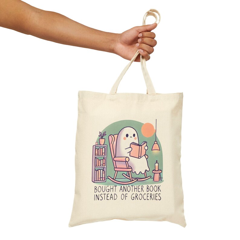 A natural-colored tote bag featuring a cute ghost reading a book with a humorous message about choosing books over groceries. The text says: Bought Another Book Instead Of Groceries. Main colors are blue, orange, white, pink and black.