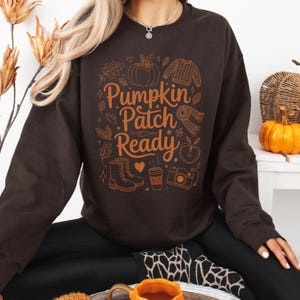 May include: A brown sweatshirt featuring the text "Pumpkin Patch Ready" in orange. The design includes illustrations of a pumpkin, sweater, scarf, apple, boots, coffee cup, and camera. This is a fall-themed item.
