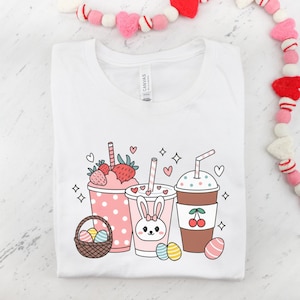 May include: White t-shirt featuring a graphic of three cartoon drinks. The drinks are a strawberry milkshake, a bunny-shaped milkshake, and a chocolate milkshake with a cherry on top. The design also includes cartoon hearts, stars, and Easter eggs.