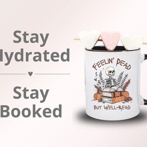A white mug with a black or pink handle featuring a skeleton reading a book, surrounded by fall leaves and pumpkins, with the text: Feelin' Dead But Well-Read. Main colors are brown and orange.