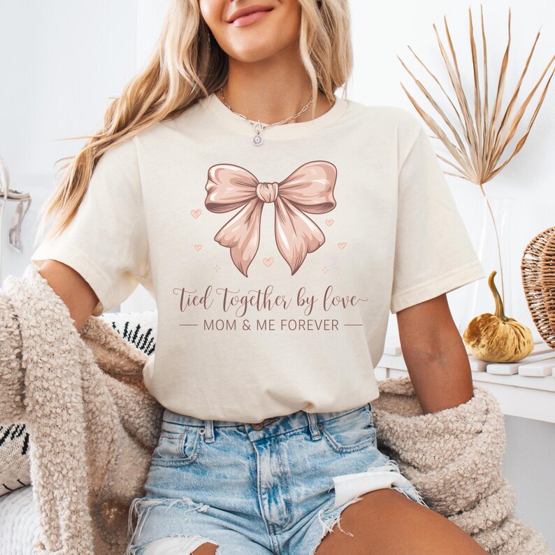 A sentimental t-shirt featuring a beautifully illustrated silk bow with the phrase: Tied Together by Love – Mom & Me Forever. Small hearts surround the design, making it a sweet and emotional tribute to the mother-daughter bond. The colors are very soft and all in pink tones.
