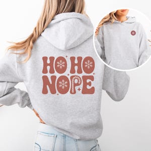 May include: A light gray hoodie with the words "HO HO NOPE" in a retro font on the back. The design includes snowflakes and a candy cane. A smaller snowflake design is on the front. The hoodie has a kangaroo pocket and a hood.