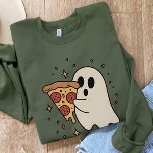 May include: Olive green sweatshirt featuring a cartoon ghost holding a slice of pizza. The pizza has pepperoni and a golden crust. The design includes stars and small circles. The sweatshirt is on a wooden surface.