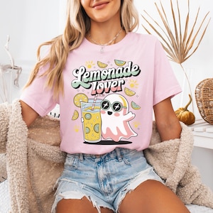 Peut inclure: T-shirt rose clair avec l'inscription "Lemonade Lover" dans une police r&eacute;tro. Le motif repr&eacute;sente un fant&ocirc;me portant des lunettes de soleil, tenant un verre de limonade, entour&eacute; de tranches de citron et de soleils.