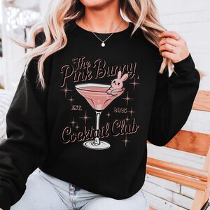 A sweatshirt featuring a pink martini glass with a bunny lounging on the rim, surrounded by vintage-style lettering that reads: The Pink Bunny Cocktail Club, Est. 2025. The main colors are pink and black.