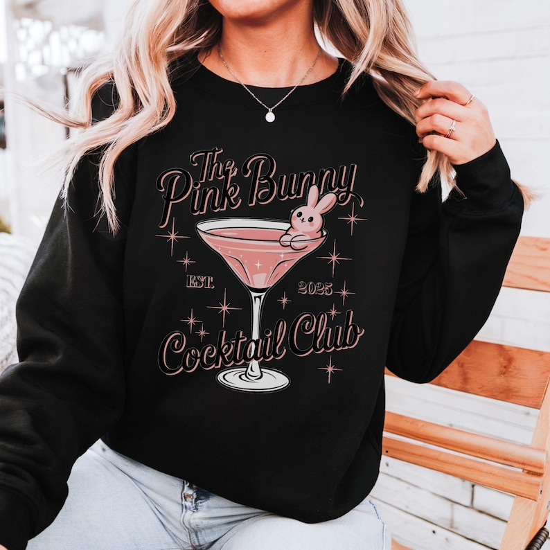 A sweatshirt featuring a pink martini glass with a bunny lounging on the rim, surrounded by vintage-style lettering that reads: The Pink Bunny Cocktail Club, Est. 2025. The main colors are pink and black.