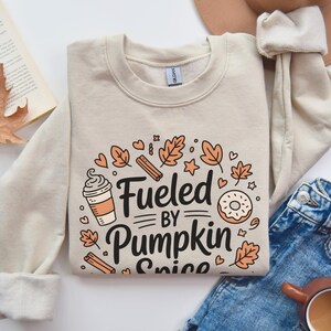 Op de afbeelding: Beige sweatshirt met de tekst "Fueled by Pumpkin Spice" in zwart, omringd door herfstthema-illustraties. Het ontwerp bevat een koffiekopje, donut, kaneelstokjes en bladeren. De sweatshirt ligt op een witte ondergrond.