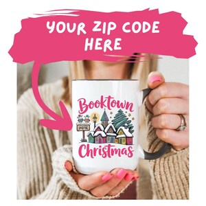 May include: White ceramic mug with a black handle featuring the words "Booktown Christmas" in pink and a winter village scene. The mug also has the text "Your Zip Code Here" in a pink speech bubble.
