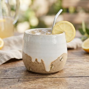 Può includere: Un bicchiere in ceramica fatto a mano con una glassa bianca su una base marrone macchiata, pieno di tè freddo e una fetta di limone. Una cannuccia di metallo è inserita nella bevanda. La tazza è su un tavolo di legno, suggerendo una bevanda rinfrescante.