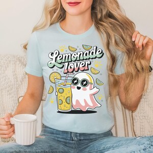 Peut inclure: T-shirt bleu clair avec l'inscription "Lemonade Lover" dans une police r&eacute;tro. Le motif repr&eacute;sente un fant&ocirc;me de dessin anim&eacute; portant des lunettes de soleil, tenant un verre de limonade avec des tranches de citron. Le t-shirt est de couleur pastel.