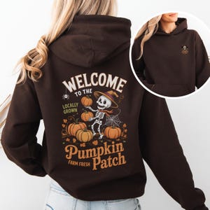 Op de afbeelding: Een donkerbruine hoodie met een Halloween-thema op de achterkant. De afbeelding toont een skelet met een hoed en sjaal, pompoenen vasthoudend, met de tekst "WELCOME TO THE PUMPKIN PATCH". Een kleinere afbeelding staat op de voorkant.