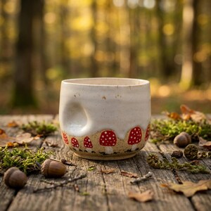 May include: A ceramic mug with a speckled off-white glaze and a thumb indentation on the side. The lower portion features a tan base decorated with a repeating pattern of red mushrooms with white spots, encircling the entire exterior surface.