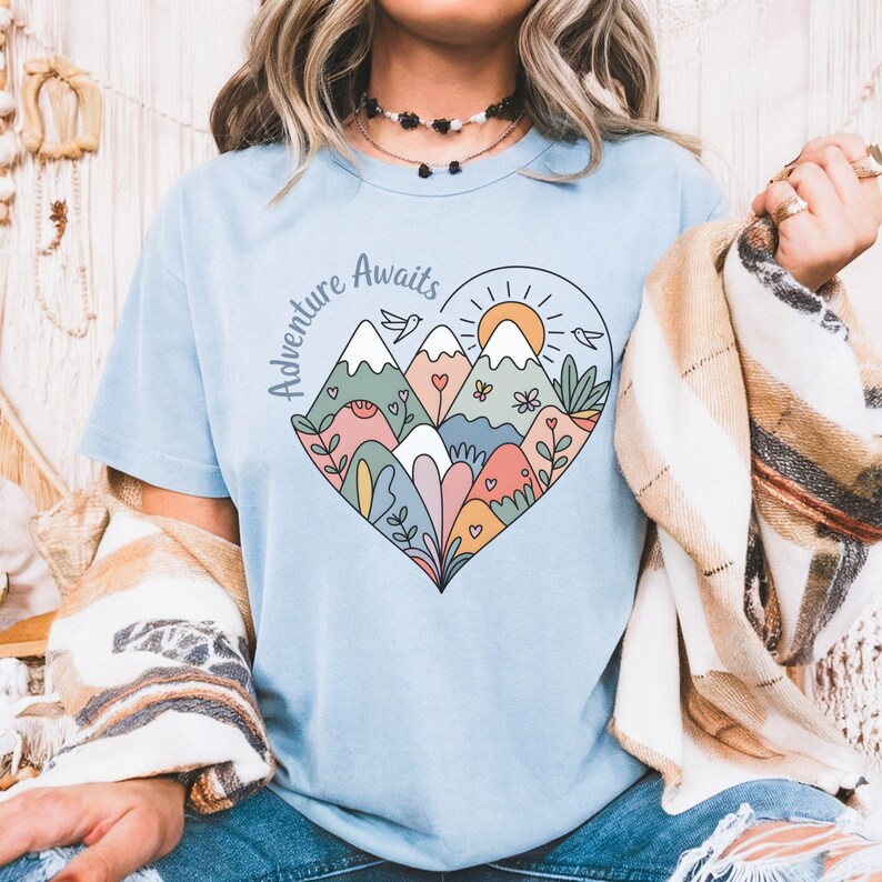 T-shirt featuring a colorful heart-shaped mountain design filled with wildflowers, sun, birds, and nature elements. Text reads “Adventure Awaits.” A cheerful outdoorsy aesthetic ideal for travel lovers, hikers, and boho nature girls.