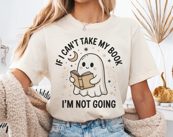 May include: Cream-colored t-shirt with the text "IF I CAN'T TAKE MY BOOK I'M NOT GOING" and a cartoon ghost reading a book. The design includes a crescent moon, stars, and constellations. The shirt is paired with blue denim shorts.