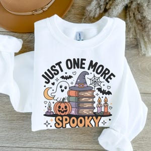 May include: White sweatshirt with a Halloween-themed graphic. The design includes a ghost, a stack of books topped with a witch hat, a jack-o'-lantern, bats, candles, and the text "JUST ONE MORE SPOOKY".