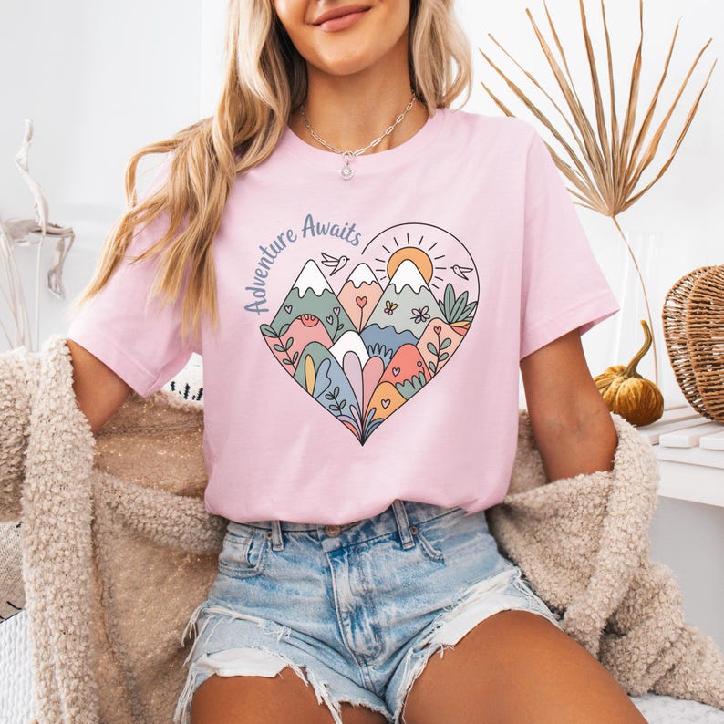 T-shirt featuring a colorful heart-shaped mountain design filled with wildflowers, sun, birds, and nature elements. Text reads “Adventure Awaits.” A cheerful outdoorsy aesthetic ideal for travel lovers, hikers, and boho nature girls.