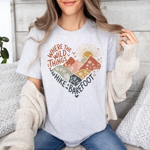 T-shirt with a heart-shaped mountain and floral design, sun rising in the background. Text reads: Where the Wild Things Hike Barefoot. The main colors are orange, green, red and yellow tones.