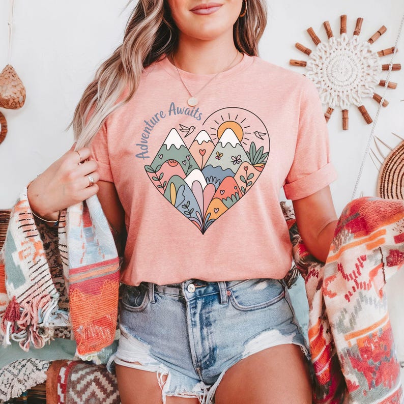 T-shirt featuring a colorful heart-shaped mountain design filled with wildflowers, sun, birds, and nature elements. Text reads “Adventure Awaits.” A cheerful outdoorsy aesthetic ideal for travel lovers, hikers, and boho nature girls.
