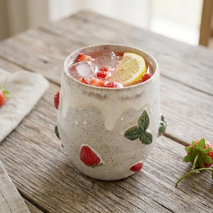 Può includere: Un bicchiere in ceramica con smalto bianco sporco maculato e un motivo a goccia color crema sul bordo. L'esterno presenta motivi tridimensionali in rilievo di fragole rosse e foglie verdi. La tazza è riempita con ghiaccio, fragole a fette e una fetta di limone.