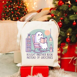 A natural-colored tote bag featuring a cute ghost reading a book with a humorous message about choosing books over groceries. The text says: Bought Another Book Instead Of Groceries. Main colors are blue, orange, white, pink and black.