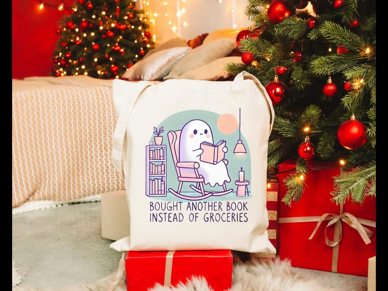 A natural-colored tote bag featuring a cute ghost reading a book with a humorous message about choosing books over groceries. The text says: Bought Another Book Instead Of Groceries. Main colors are blue, orange, white, pink and black.