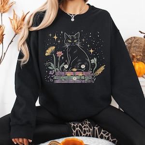 Op de afbeelding: Zwart sweatshirt met een illustratie van een zwarte kat met gele ogen, zittend op boeken. Het ontwerp bevat kleurrijke bloemen, sterren en een hemelse achtergrond. Een comfortabel en stijlvol kledingstuk voor kattenliefhebbers.