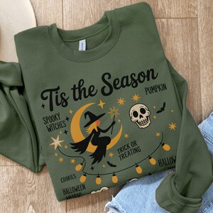 Könnte beinhalten: Olivgrünes Sweatshirt mit einem Halloween-Motiv. Das Design zeigt eine Hexe auf einem Mond, einen Totenkopf, Sterne und den Text "Tis the Season". Zusätzlicher Text: "Spooky Witches", "Pumpkin", "Trick or Treating", "Cookies", "Halloween" und "Music".