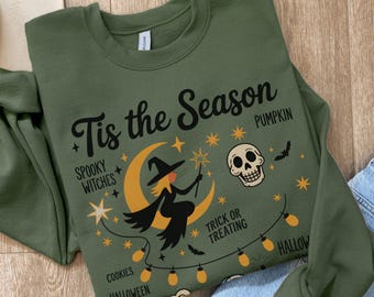 May include: Olive green sweatshirt with a Halloween-themed graphic. The design includes a witch on a crescent moon, a skull, stars, and the text "Tis the Season". Additional text includes "Spooky Witches", "Pumpkin", "Trick or Treating", "Cookies", "Halloween", and "Music".