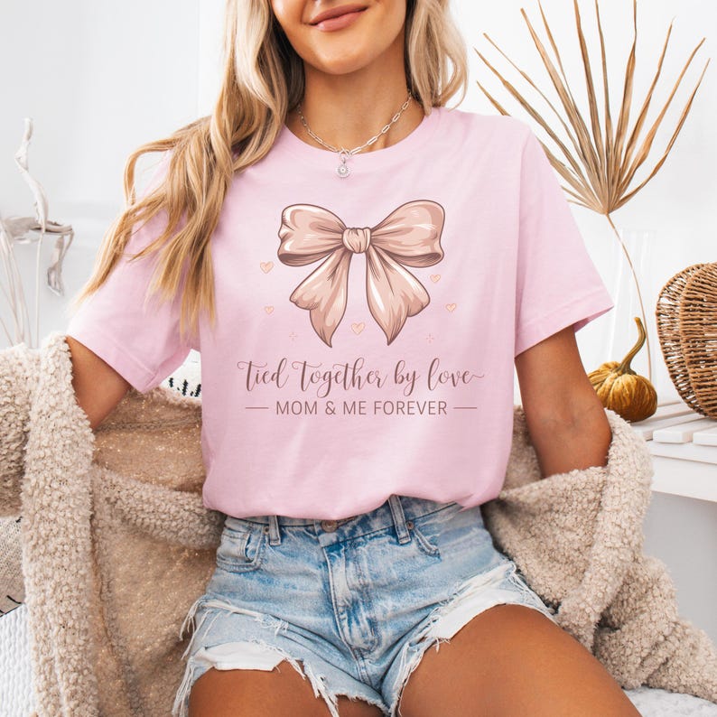 A sentimental t-shirt featuring a beautifully illustrated silk bow with the phrase: Tied Together by Love – Mom & Me Forever. Small hearts surround the design, making it a sweet and emotional tribute to the mother-daughter bond. The colors are very soft and all in pink tones.