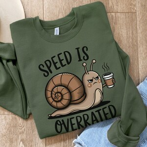 May include: Olive green sweatshirt with the text "SPEED IS OVERRATED" and a cartoon snail holding a coffee cup. The snail has an angry expression. The sweatshirt is folded on a wooden surface.