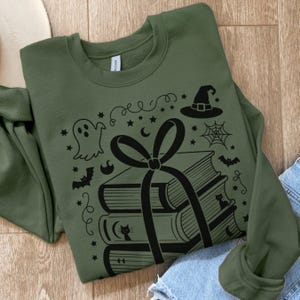 May include: Olive green sweatshirt with a black graphic of stacked books tied with a bow, and Halloween-themed illustrations including a ghost, witch hat, bats, and spiderweb. The design is centered on the front of the long-sleeved top.