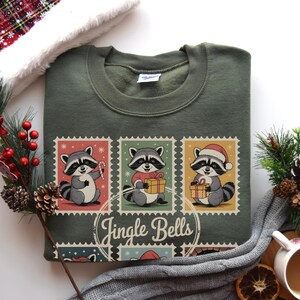 Könnte beinhalten: Olivgrünes Sweatshirt mit einem festlichen Design. Es zeigt drei Waschbären auf Briefmarken-Grafiken, jeder mit einem Urlaubsartikel. Der Text "Jingle Bells" steht darunter. Ein gemütliches Kleidungsstück für die Weihnachtszeit.