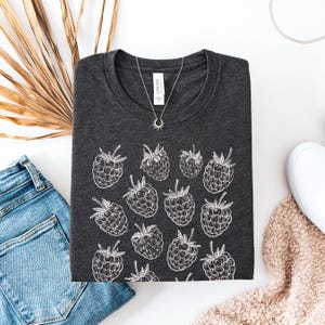 Könnte beinhalten: Ein dunkelgraues T-Shirt mit einem weißen Druck von mehreren Himbeeren. Das Shirt ist gefaltet und liegt flach auf einer weißen Oberfläche.